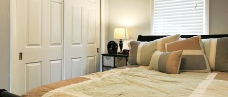 A bedroom with a bed, a nightstand, a lamp, a clock, and a window with blinds.