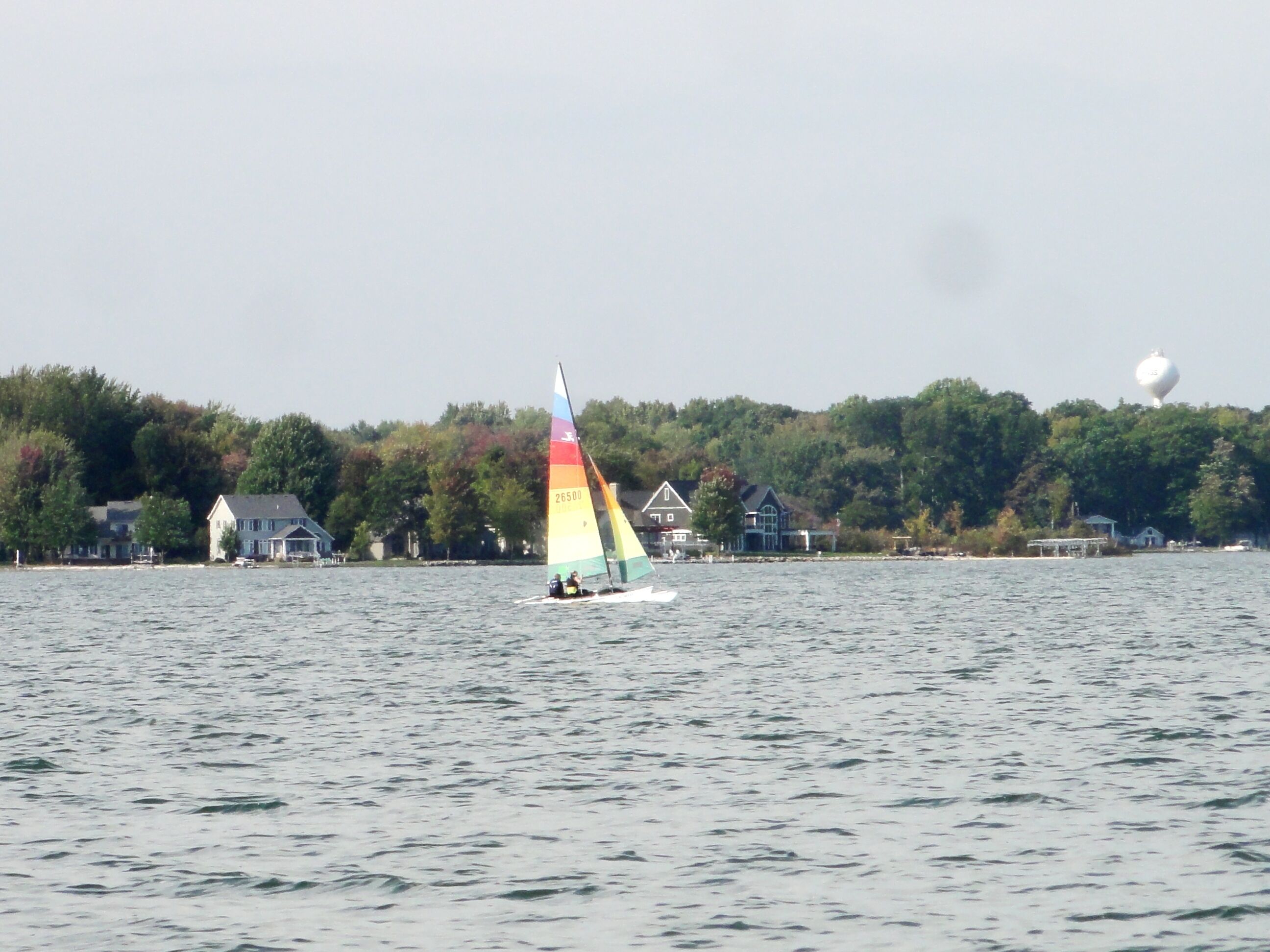 A sailboat on a lake with houses and trees in the background.