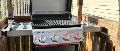 Una parrilla de gas portátil con cuatro quemadores y un estante lateral, ubicada sobre una terraza de madera.