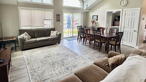 A spacious living room with a ceiling fan, a dining area with a table and chairs, a sofa, and a large window. - Luxury Gun Lake 4 Bed 2 Bath Home W/paddleboards/kayaks, Free Wi-fi (Wayland)