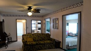 Un salon avec un ventilateur de plafond, un canapé, une table basse et une bibliothèque. Une porte donne accès à une autre pièce.