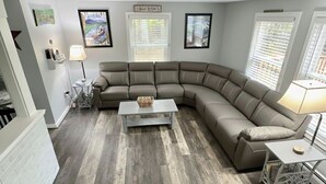 Sala de estar com sofá seccional, mesa de centro e mesa lateral. Quadros emoldurados nas paredes e janela com persianas.