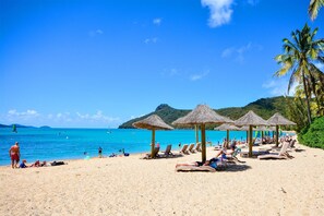 A beach with sun loungers and thatched umbrellas, a clear blue sea, and a mountainous backdrop.