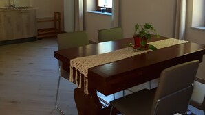 A dining area with a wooden table, chairs, and a plant. There is a kitchen area with wooden cabinets and a window. - B&B da Morena (Minucciano)