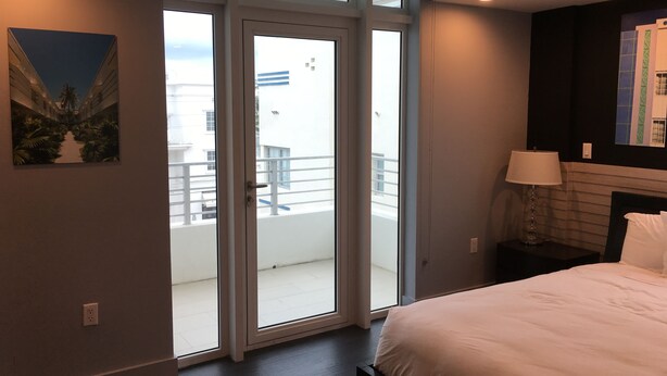 Una habitación de hotel moderna con una cama grande, una mesita de noche, una lámpara y un ventanal corredero que da a un balcón con vistas a edificios y palmeras.