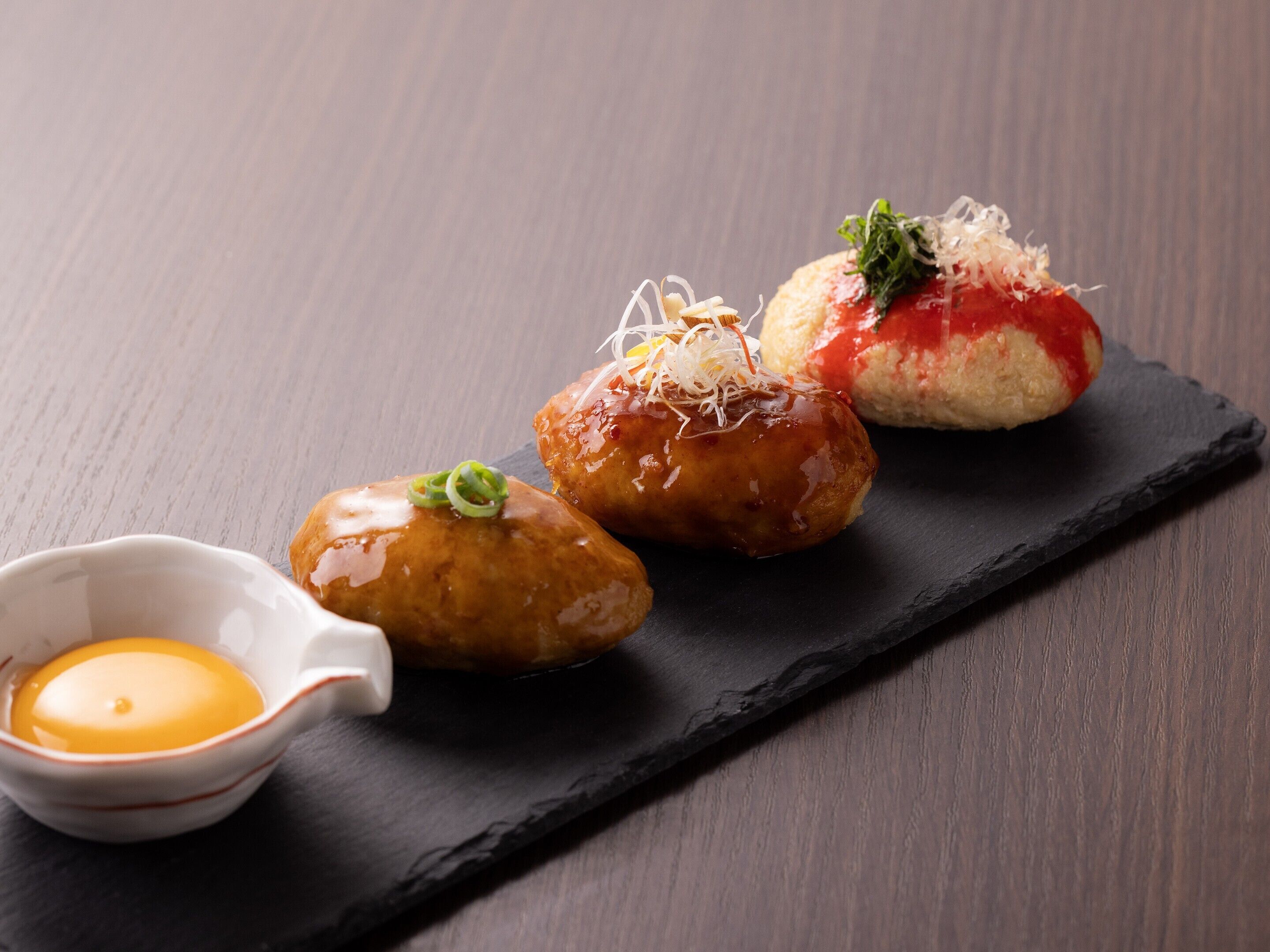 Three food items on a slate board with a small bowl of sauce.