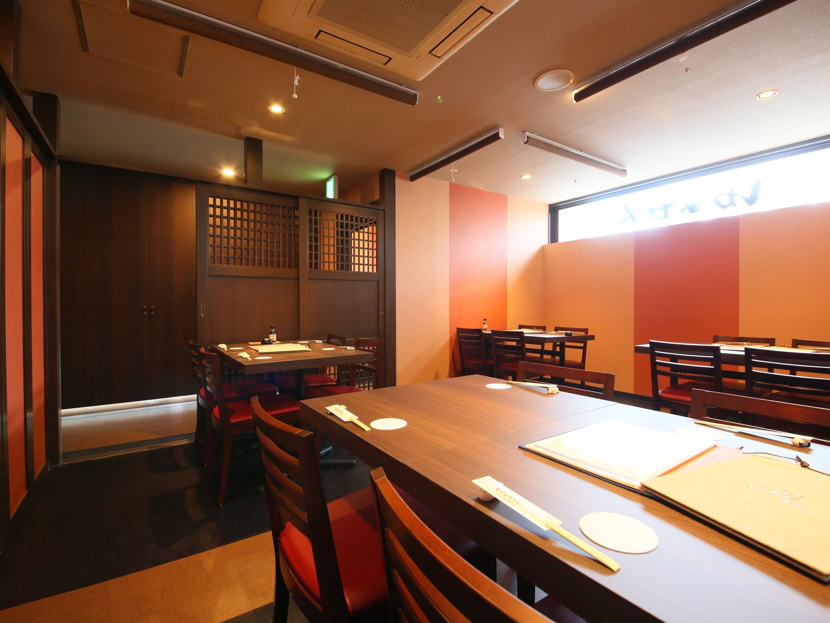 A restaurant with wooden tables and chairs, a high ceiling with recessed lighting, and a large window with a view of the street.
