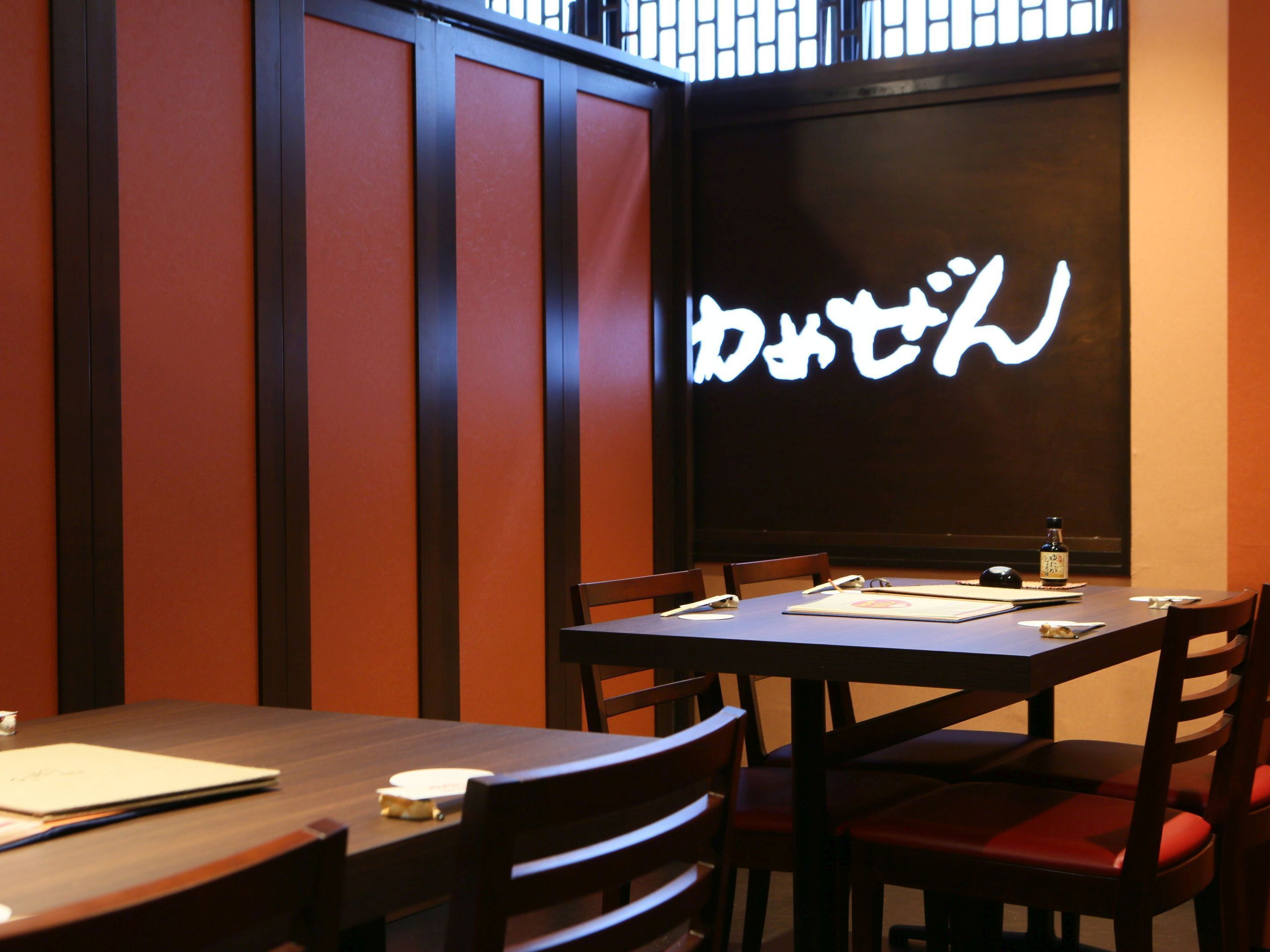 Ein Restaurantinneres mit Holztischen und einer dunklen Wand, die mit beleuchteten japanischen Schriftzeichen versehen ist.