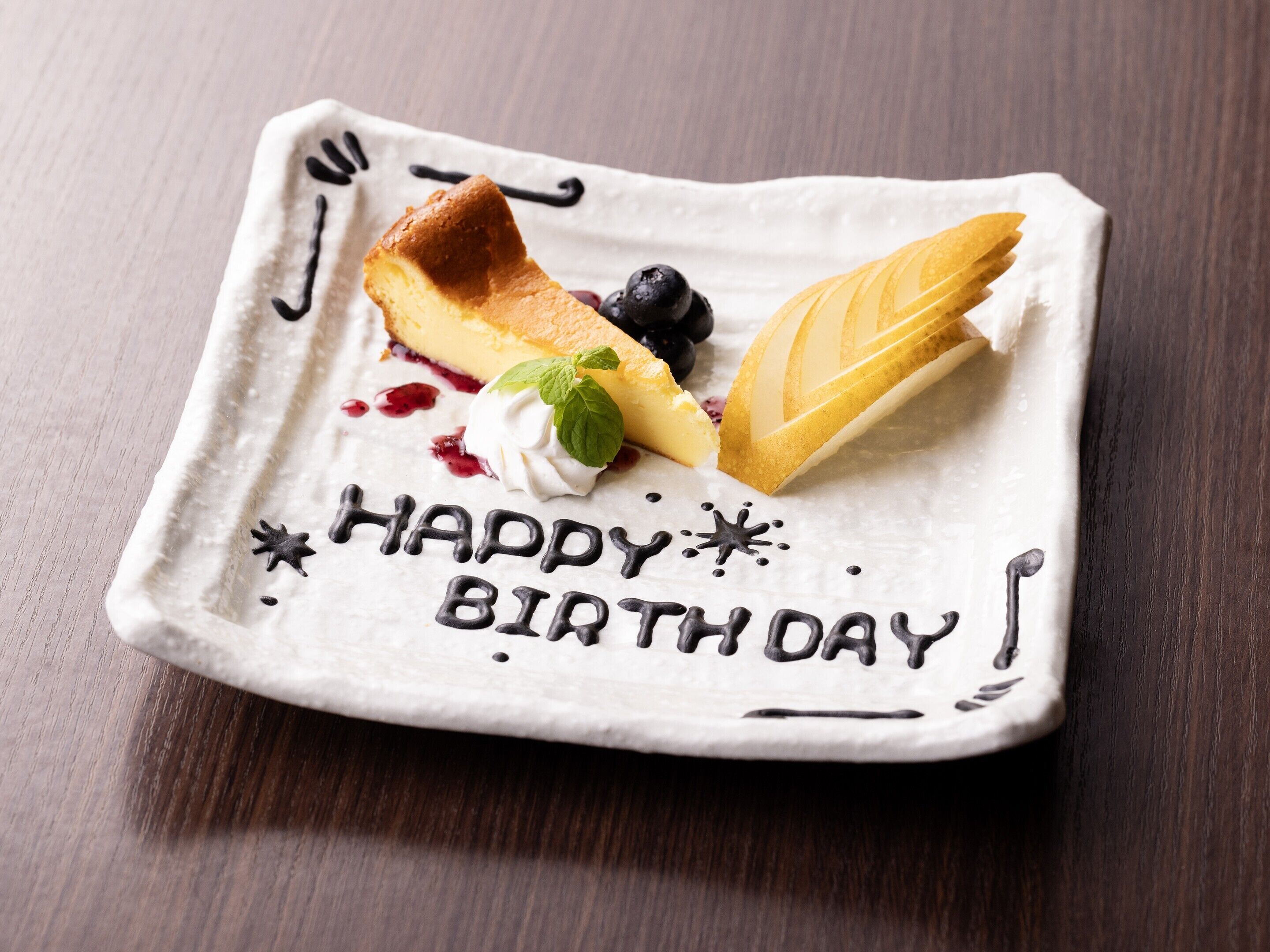 A dessert plate with a slice of cheesecake, blueberries, and a leaf-shaped pastry on a white plate with "HAPPY BIRTH DAY" written in chocolate.