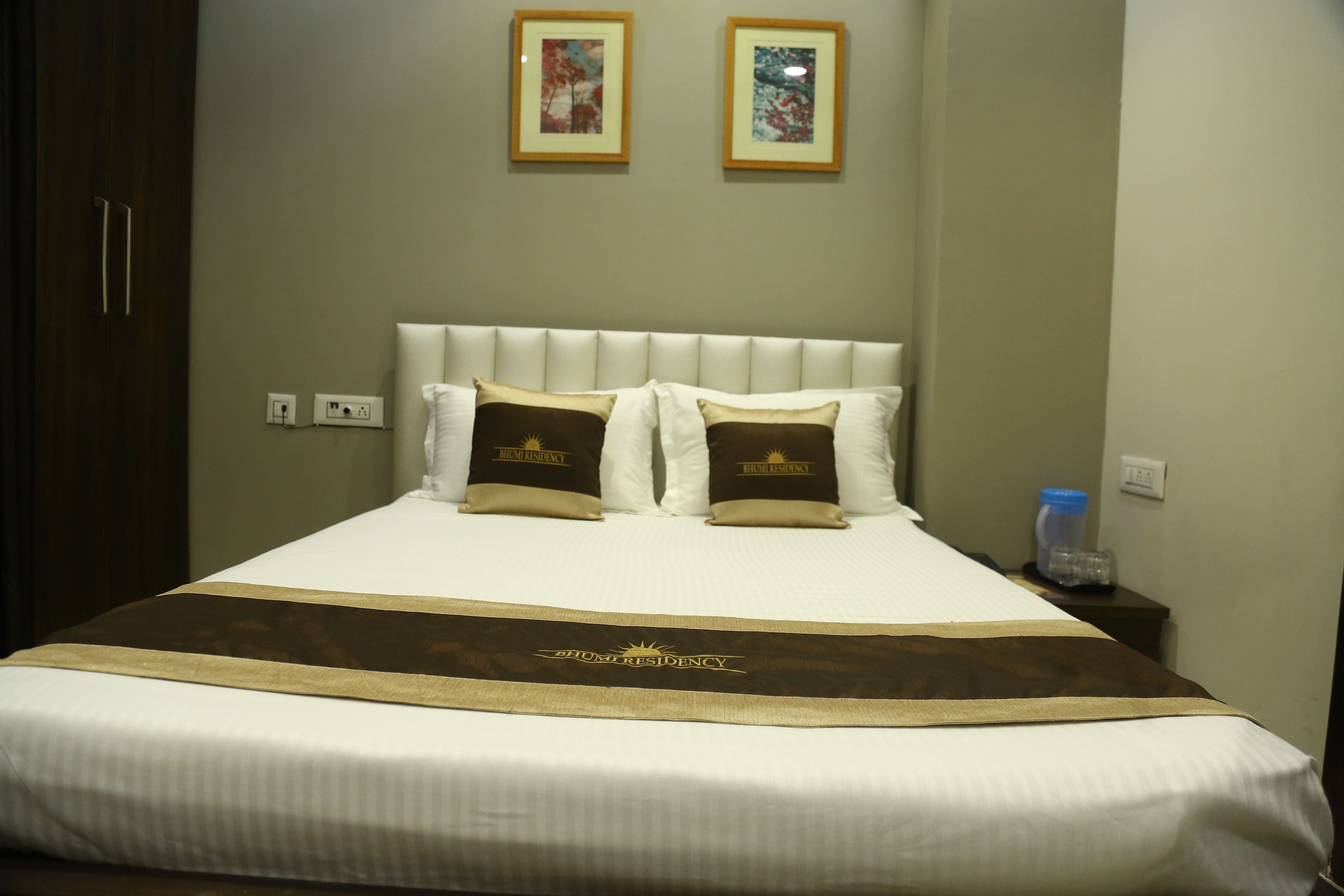 A hotel room with a large bed, a headboard, two framed pictures on the wall, and a bedside table with a water bottle.
