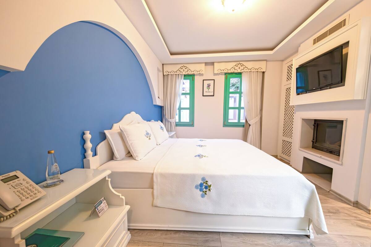 Un dormitorio con una cama, televisión, teléfono y una mesita con un libro y un vaso.