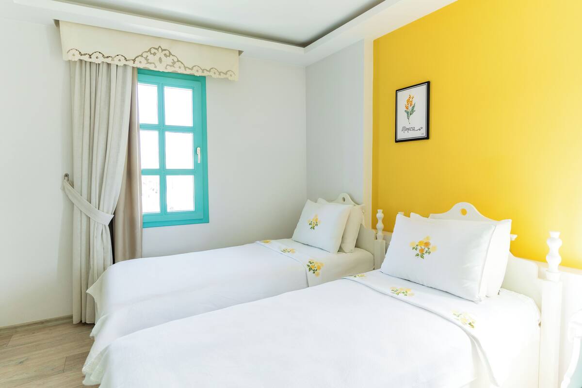 Una habitación con dos camas, una ventana con cortinas, una pared amarilla y un cuadro enmarcado.
