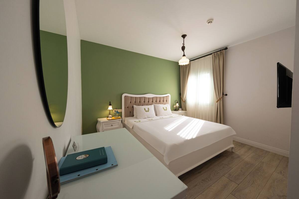 Habitación de hotel con cama, escritorio, espejo, mesita de noche y ventana con cortinas.