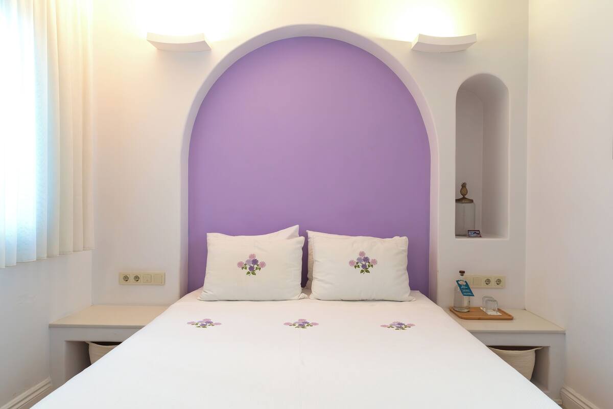 Un dormitorio con un arco morado sobre la cama, ropa de cama blanca y una mesita de noche pequeña con una lámpara y una caja de pañuelos.