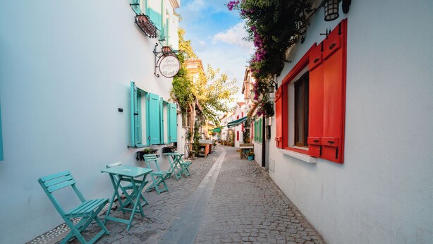Una estrecha calle adoquinada con sillas y mesas turquesas, paredes blancas y contraventanas rojas.