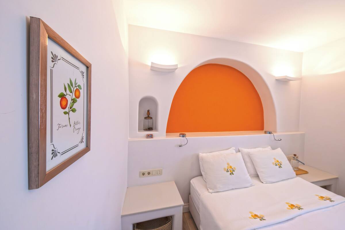 Un dormitorio con una cama, mesitas de noche blancas, un cuadro enmarcado, una pared naranja de acento y un pequeño estante con un recipiente de vidrio.