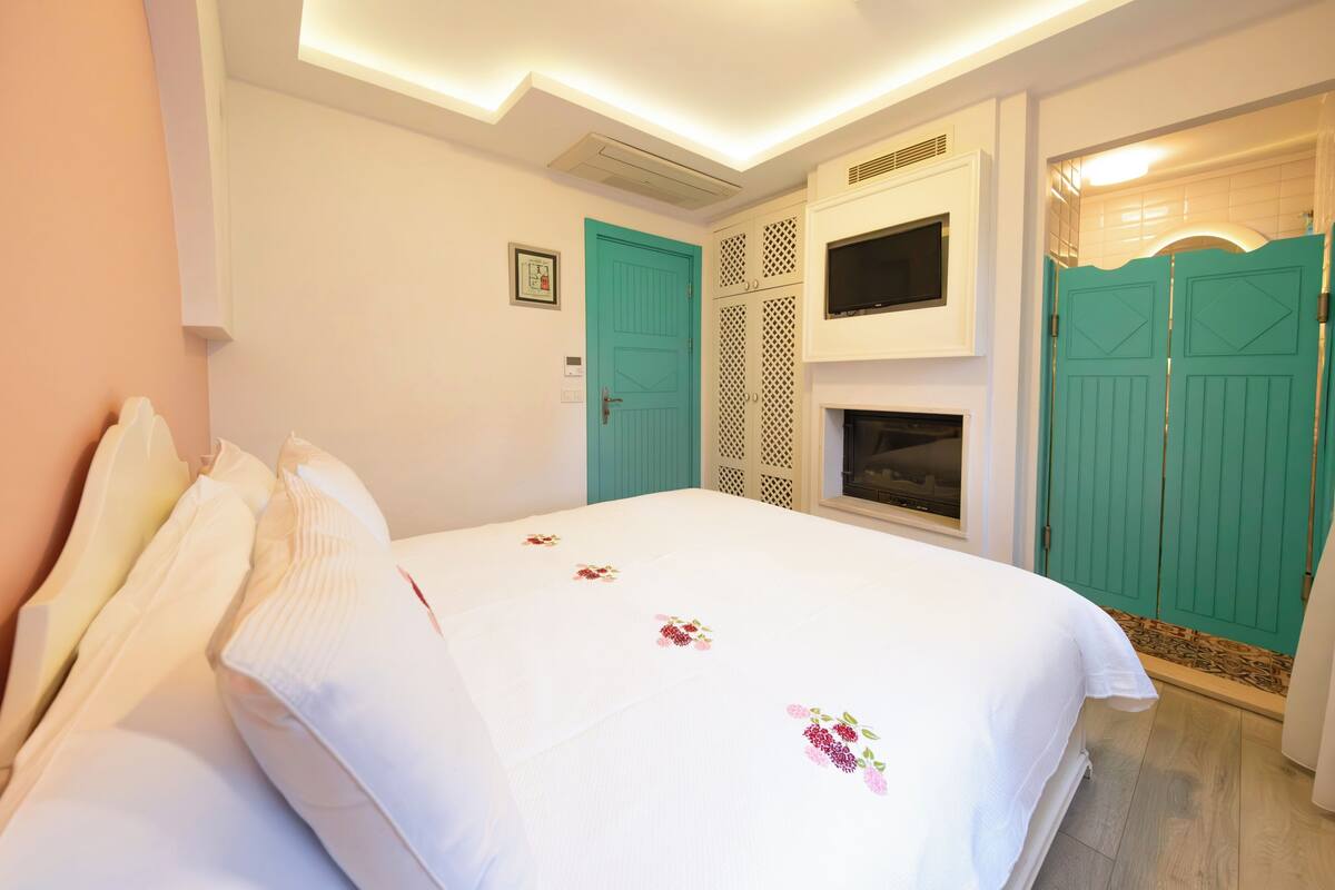 Una cama bien hecha con colcha estampada, un televisor montado encima y puertas de color verde azulado.