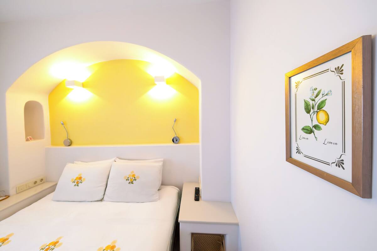 Habitación de hotel con cama, mesitas de noche, un cuadro enmarcado con motivos botánicos y una lámpara de techo con forma curva.