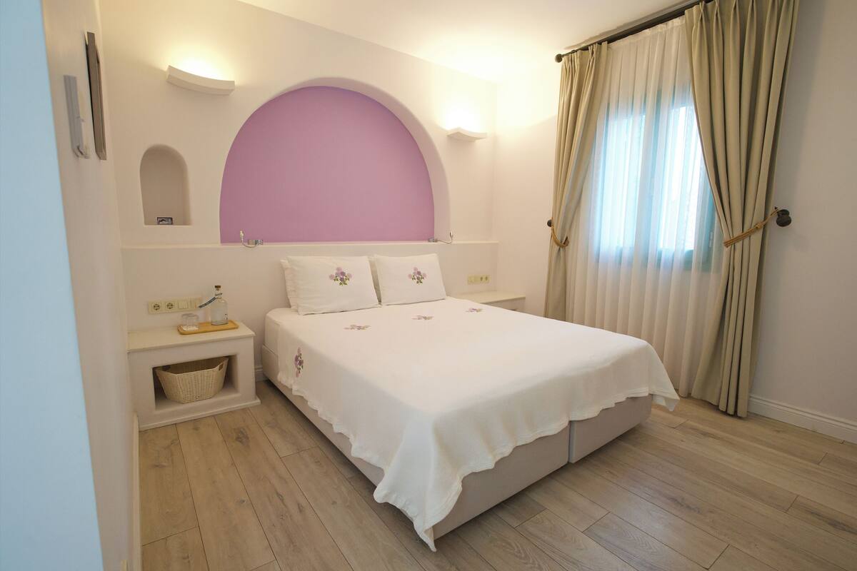 Un dormitorio con cama, mesitas de noche, estante en la pared y ventana con cortinas.