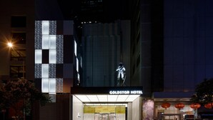 L’extérieur de l’hôtel Goldstar, avec son enseigne lumineuse bien visible, sa grande façade vitrée et son parking rempli de voitures.