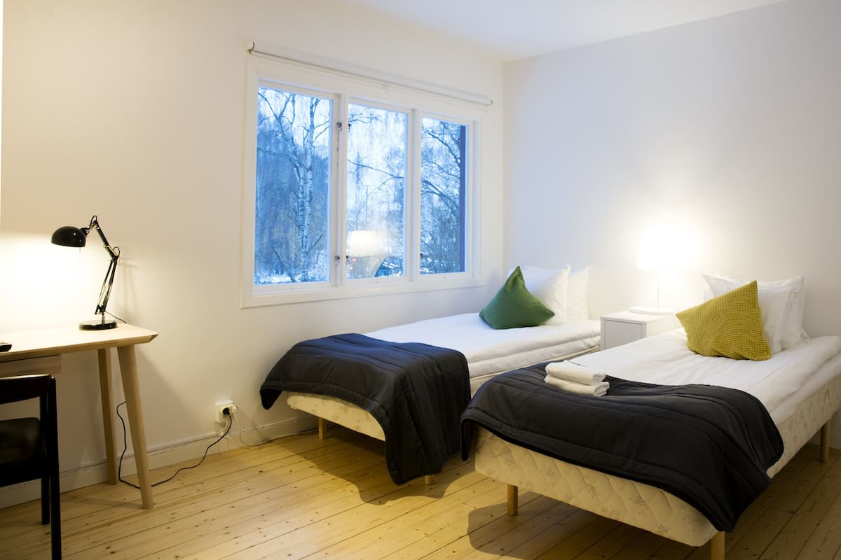 Una habitación con dos camas individuales, un escritorio con lámpara, una ventana con vistas a árboles y un enchufe de pared.