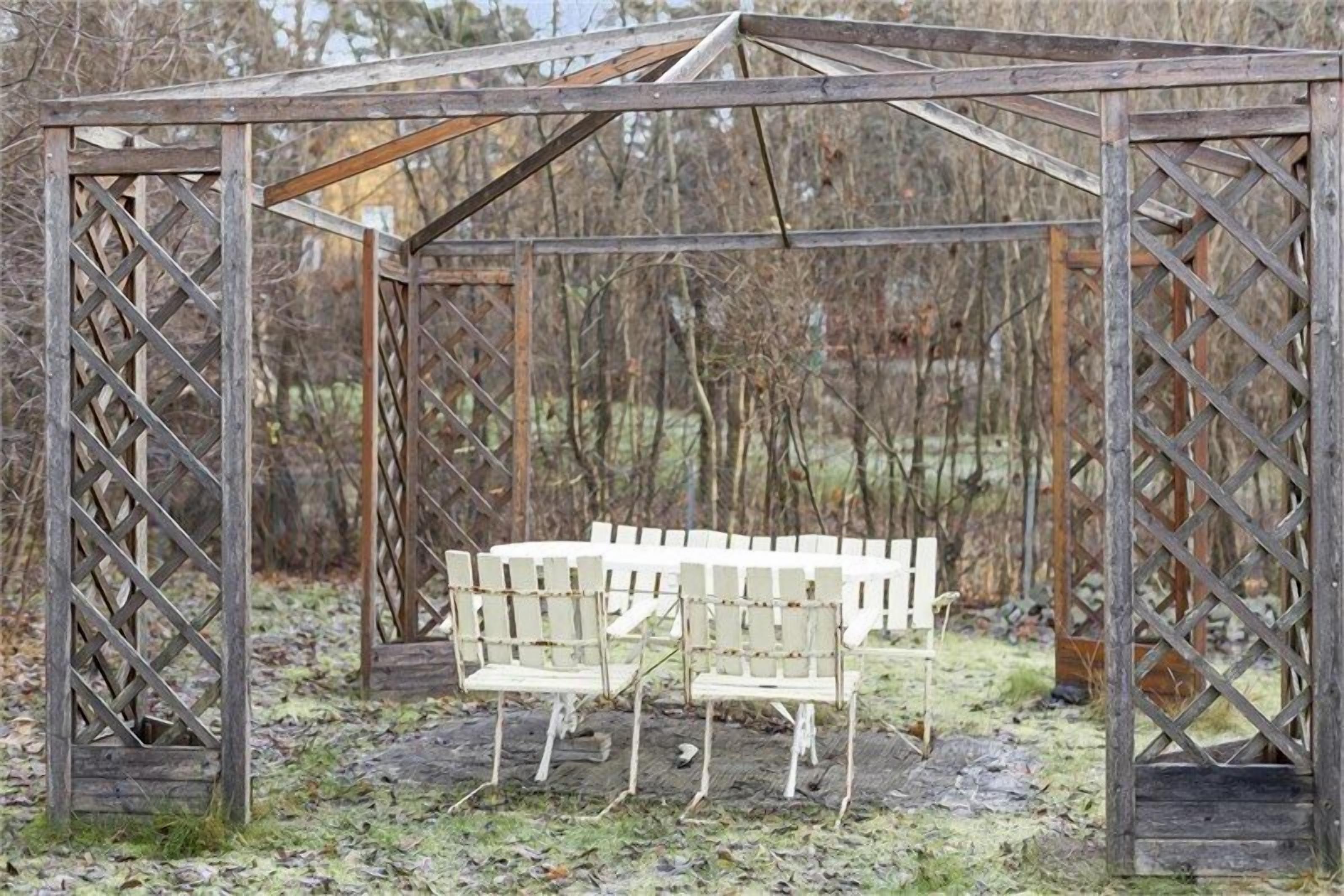 Een houten prieel met een spijlenstructuur en twee witte stoelen.