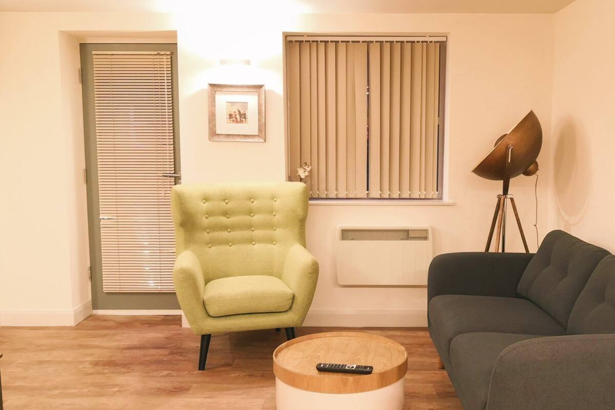 A living room with a green armchair, a dark sofa, a round coffee table with a remote control, a floor lamp, and a radiator.