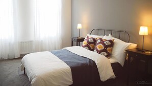 Quarto com cama, duas mesas de cabeceira, um abajur em cada mesa e janela com cortinas finas.