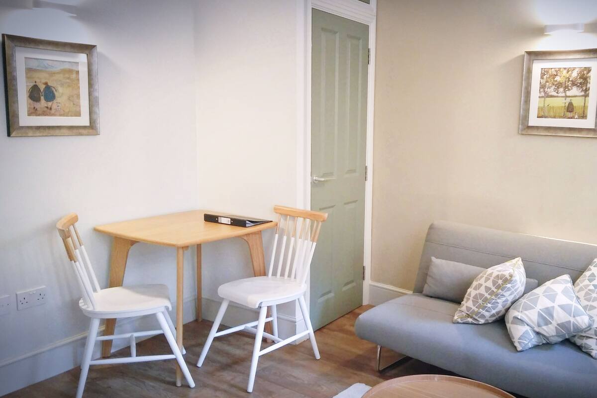A room with a wooden table and two white chairs, a grey sofa with patterned cushions, and two framed pictures on the walls.