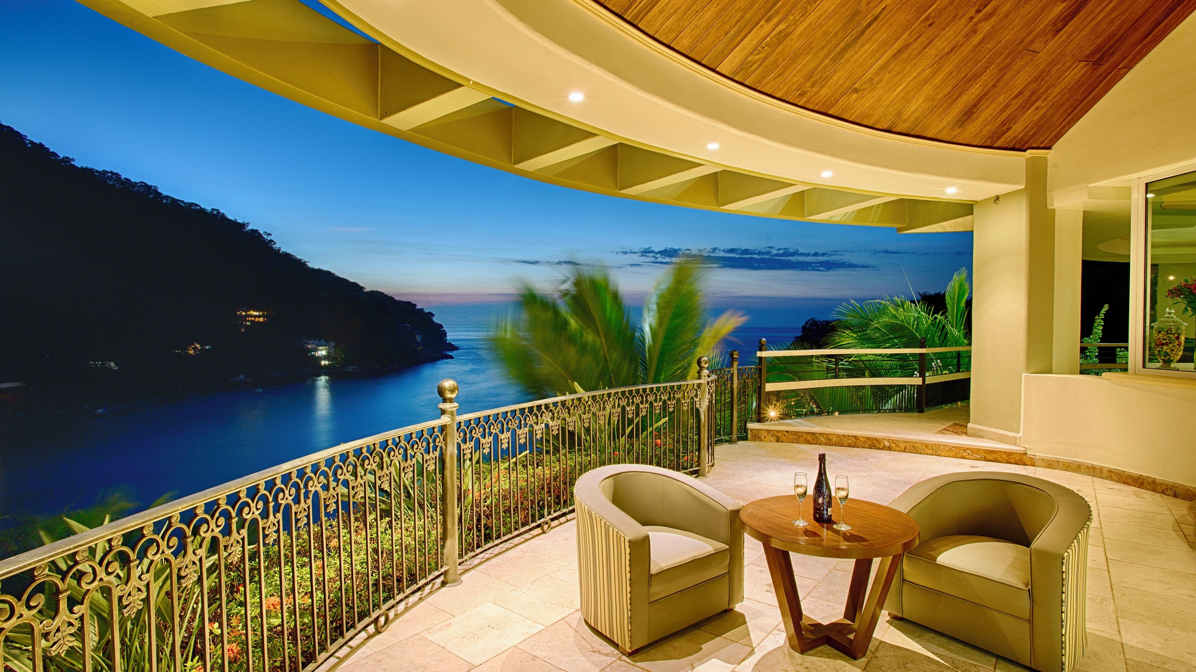Un balcon donnant sur un paysage côtier, deux fauteuils, une petite table ronde avec une bouteille et des verres, et une balustrade décorative.
