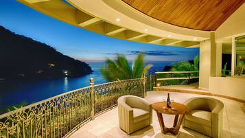 Un balcon donnant sur un paysage côtier, deux fauteuils, une petite table ronde avec une bouteille et des verres, et une balustrade décorative.