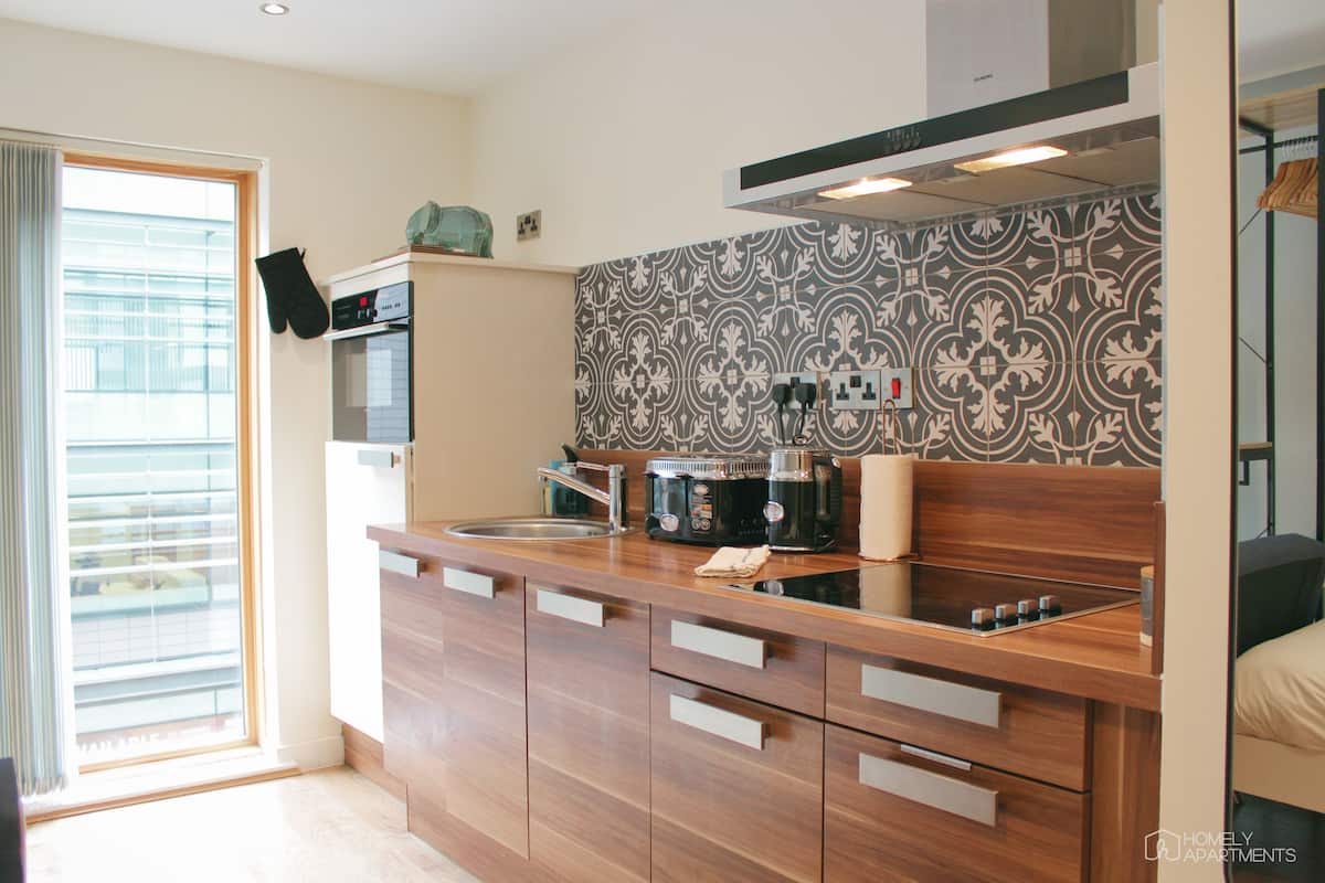 A modern kitchen with a built-in oven, a coffee maker, and a microwave. There is a window with blinds on the left side.