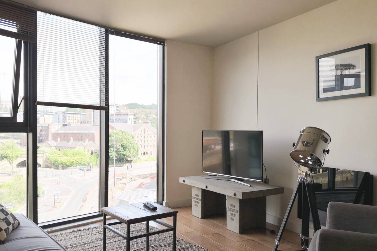 A modern living room with a large window, a television on a minimalist stand, a guitar stand, and a framed picture on the wall.