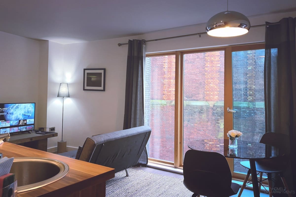 A modern living room with a television, a sofa, a dining table, and a large window with a view of a brick building.