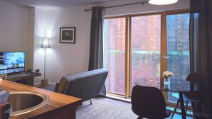 A modern living room with a television, a sofa, a dining table, and a large window with a view of a brick building. - Homely Serviced Apartments - Blonk St (Sheffield)