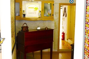 A room with a wooden cabinet, a red bench, a wall-mounted shelf with decorative items, and a fire extinguisher in the hallway. - Lilyum Alaçatı Butik Otel (Cesme)