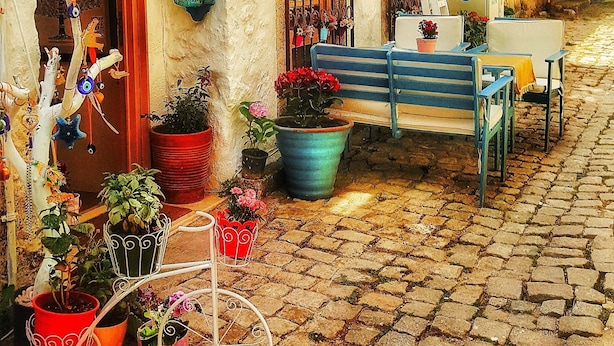 A quaint cobblestone street with a rustic building featuring a blue bench, potted plants, and a sign reading "Lilyum." - Lilyum Alaçatı Butik Otel (Cesme)