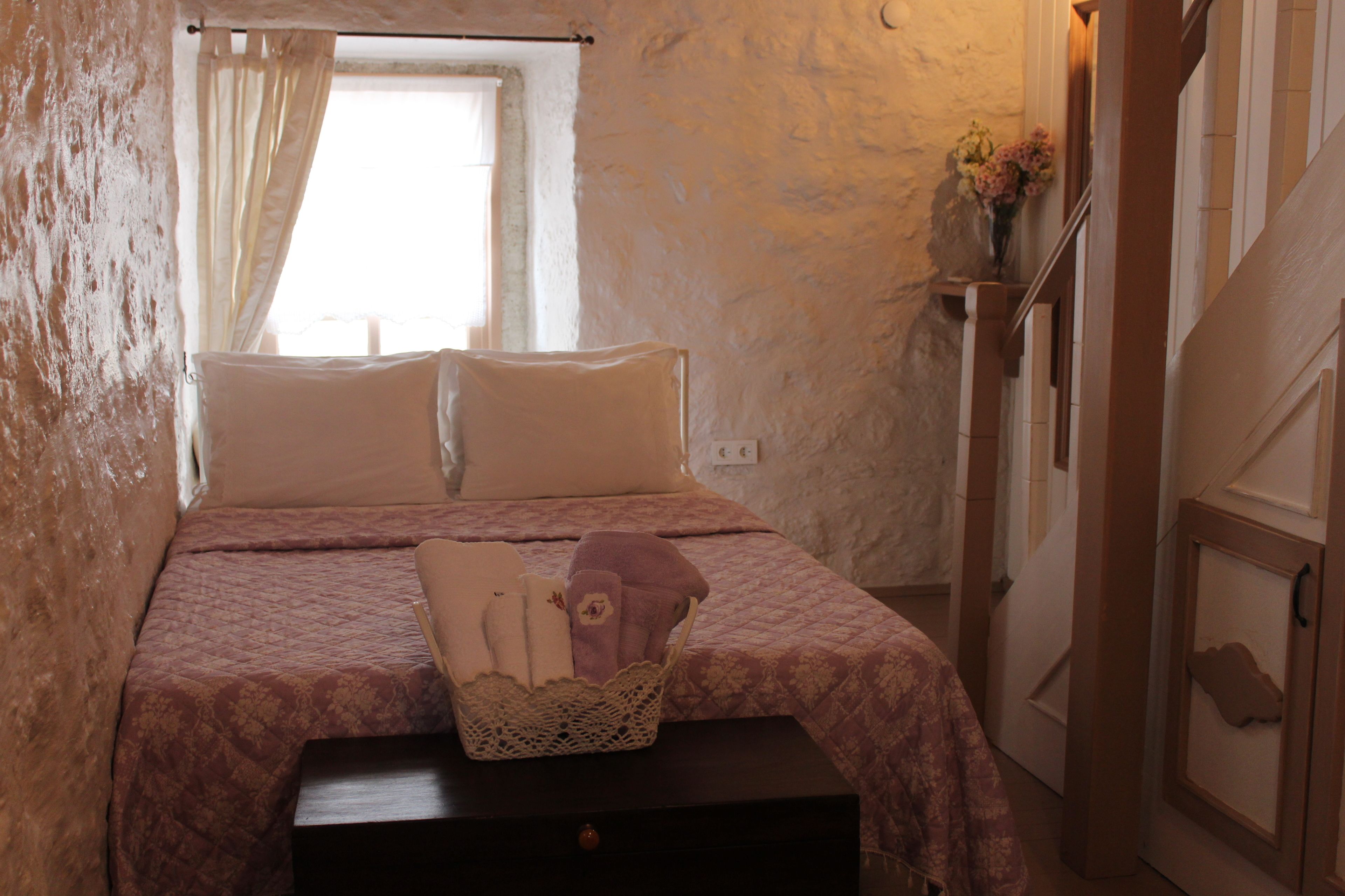 Un dormitorio con cama, almohadas, una mesita de noche, una cesta y una ventana con cortinas.