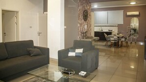 A modern living room with a grey sofa, a glass coffee table, and a grey armchair. There is a kitchen area in the background with a dining table and chairs. - Sapphire Court (Accra)