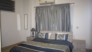A bedroom with a bed, nightstands, a lamp, and a window with blinds. - Sapphire Court (Accra)