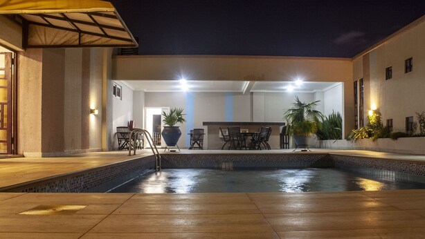 An outdoor pool area with a tiled edge, a wooden deck, and a bar area with chairs and potted plants. - Sapphire Court (Accra)