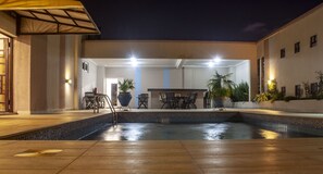 An outdoor pool area with a tiled edge, a wooden deck, and a bar area with chairs and potted plants.