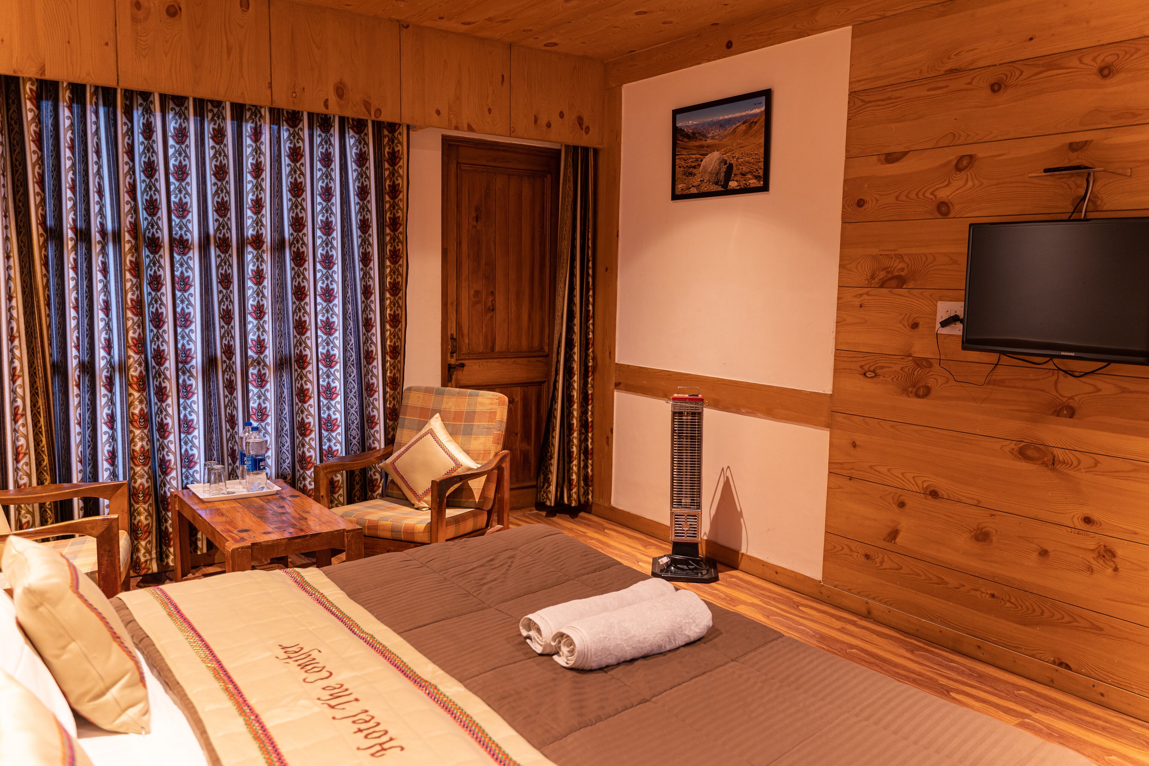 A wooden-paneled room with a bed, a chair, a small table, a heater, and a TV.