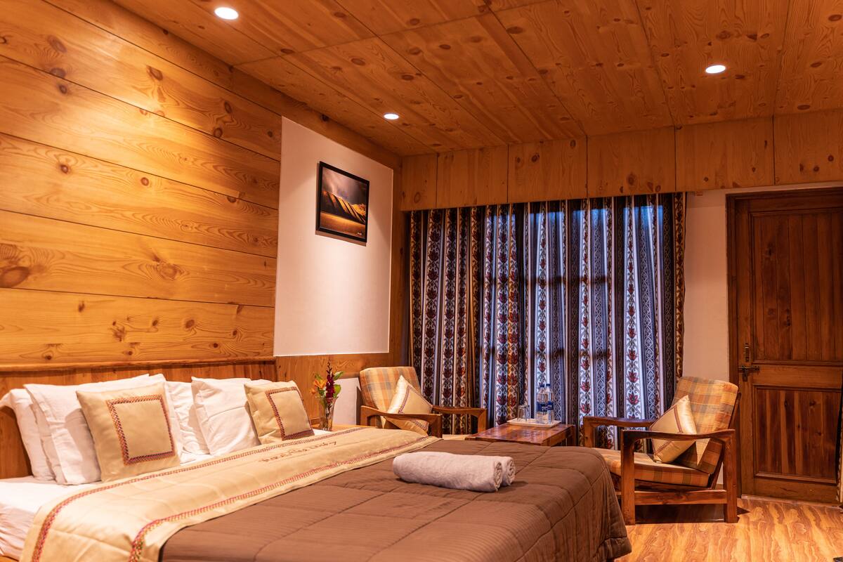 Una habitación con paneles de madera, una cama grande, dos sillones, una mesita y una ventana con cortinas estampadas.