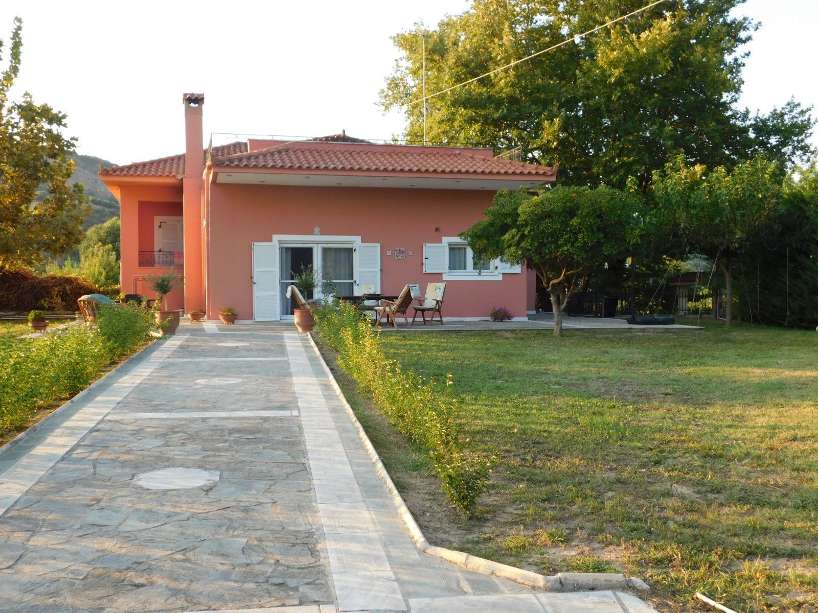 A house with a stone pathway leading to the entrance, a small patio with chairs, and a garden with trees and shrubs.