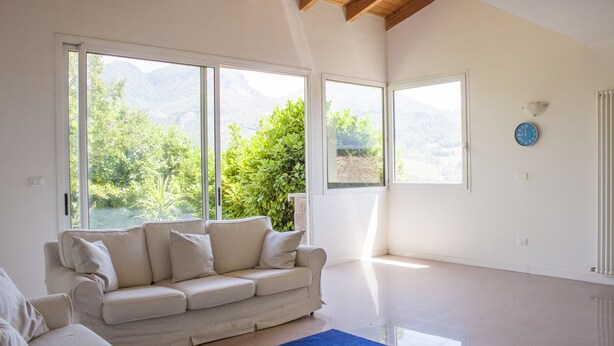 A spacious living room with a large white sofa, a blue rug, and large windows offering a mountain view. - Casa Bella Il Castello (Vergato)