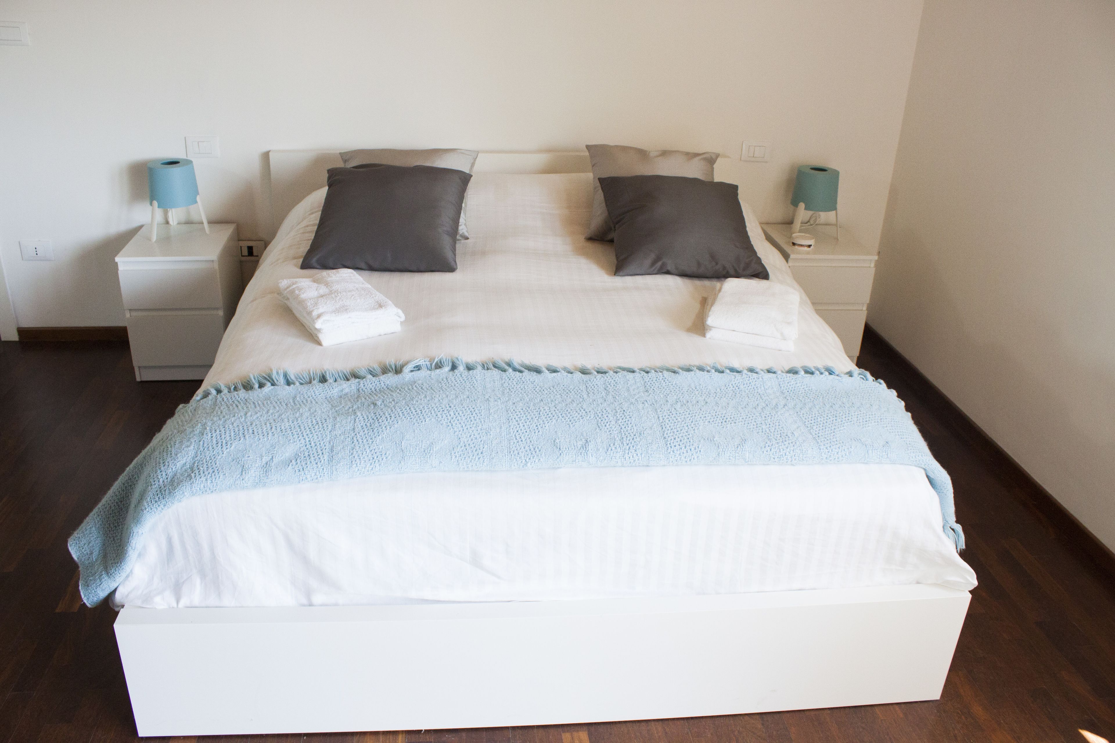 Cama bem arrumada com manta branca e azul clara, duas almofadas escuras e cabeceira branca. Há duas mesas de cabeceira com luminárias e uma gavetinha.