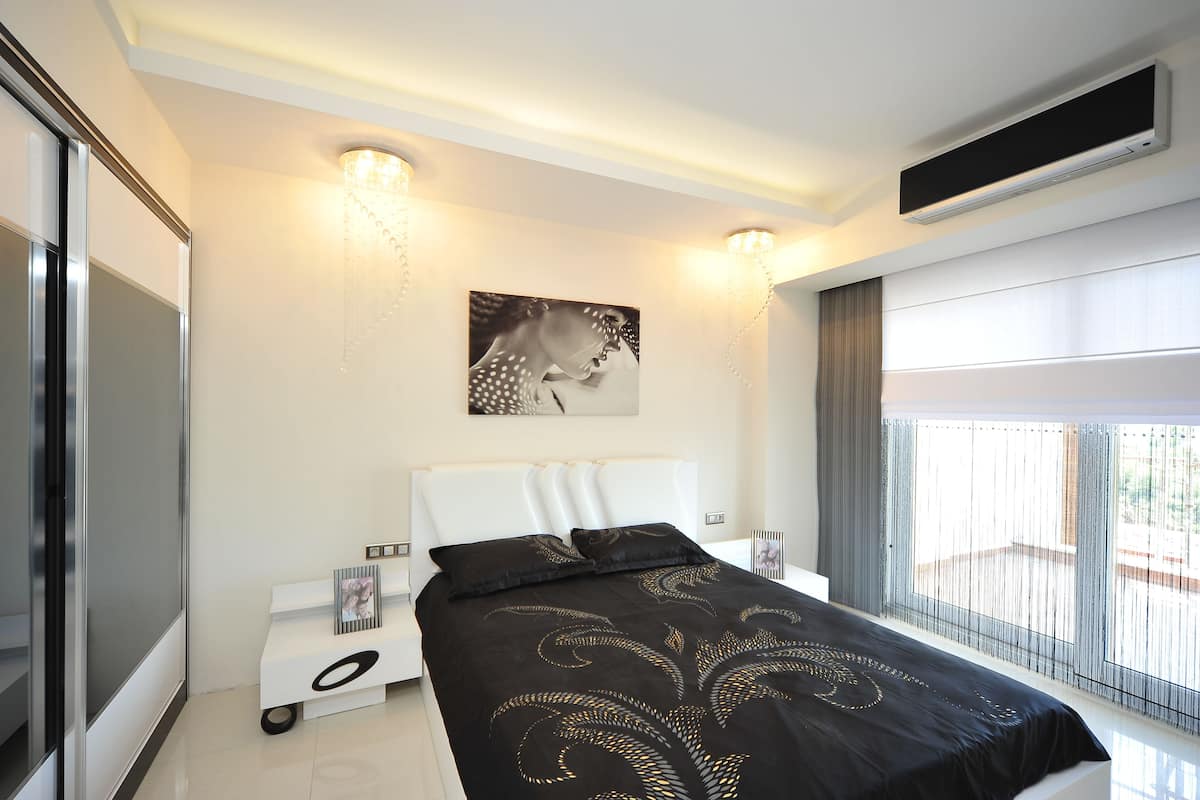 A modern bedroom with a large bed, a bedside table with a lamp, a wall-mounted TV, and a large window with blinds.