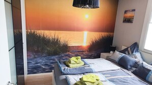 A bedroom with a mural of a sunset over water, a bed with blue and white bedding, and a green towel on the bed. - The cottage in Taddigshörn - nordseereise-net (Esens)