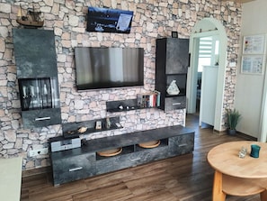 A modern living room with a stone wall, a flat-screen TV, built-in shelves, and a wooden floor. - The cottage in Taddigshörn - nordseereise-net (Esens)